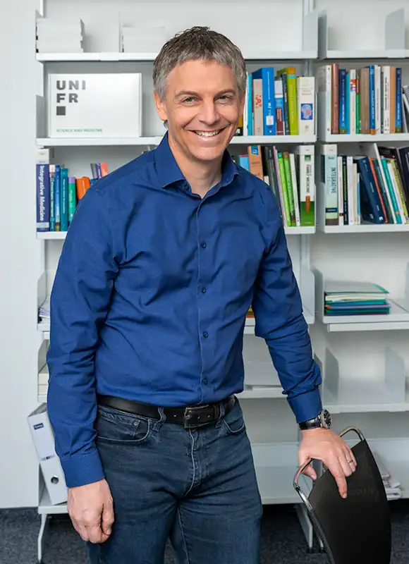 Portrait des vor einem Büchergestell posierenden Dr. med. Pierre-Yves Rodondi, ordentlicher Professor, Universität Freiburg, in blauem Hemd